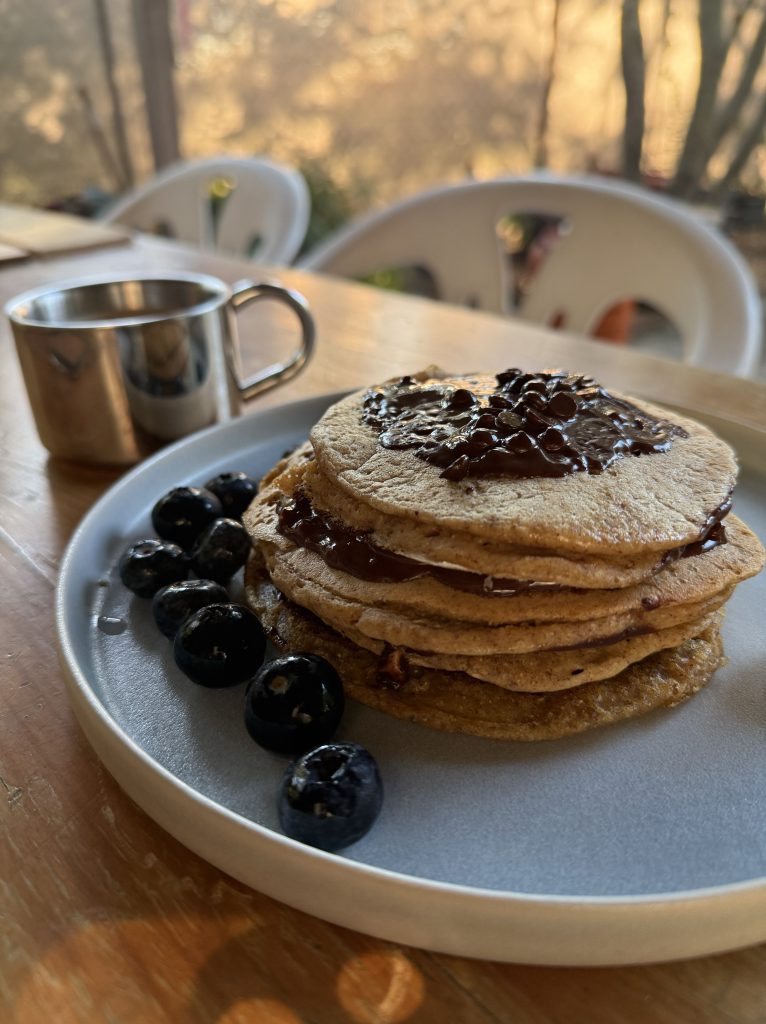 Torre de pancakes con chocolate y arándanos en un plato, junto a una pequeña jarra de jarabe.