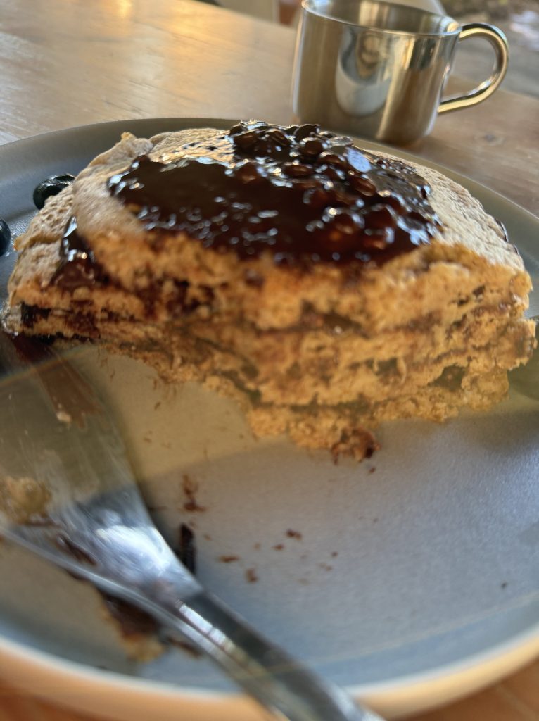 Un pancake de avena cubierto con jarabe de chocolate, acompañado de un tenedor y una taza metálica en un plato.